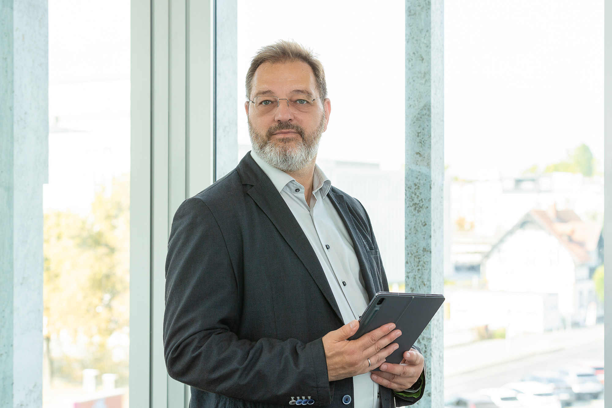 Ein älterer Mann mit grauem Bart im dunklen Anzug hält ein Tablet und blickt ruhig